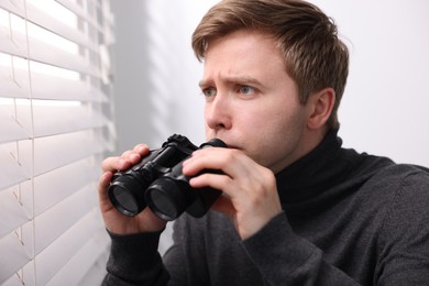 Photo of Paranoia. Worried man with binoculars near window indoors