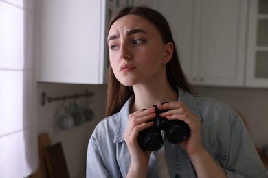 Photo of Paranoia. Worried woman with binoculars in kitchen, space for text