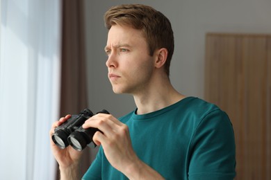 Photo of Paranoia. Worried man with binoculars near window indoors