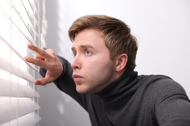 Photo of Paranoia. Worried man looking through window blinds indoors
