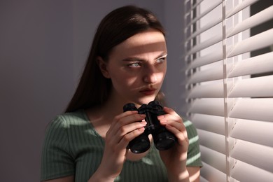 Photo of Paranoia. Worried woman with binoculars near window indoors