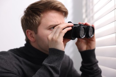 Photo of Paranoia. Worried man with binoculars near window indoors