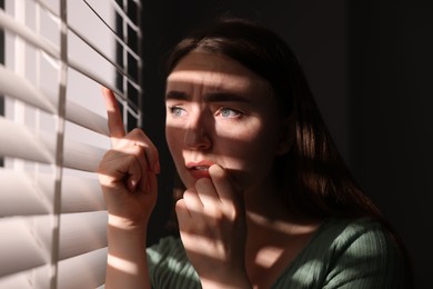 Photo of Paranoia. Worried woman looking through window blinds indoors