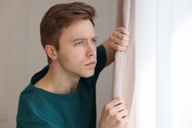 Photo of Paranoia. Worried man looking out window indoors