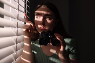 Photo of Paranoia. Worried woman with binoculars near window indoors