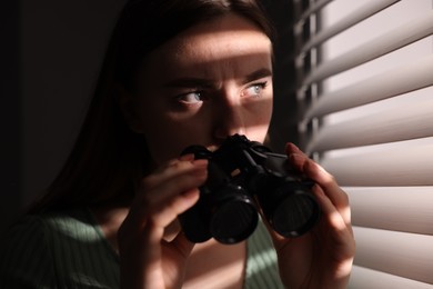 Photo of Paranoia. Worried woman with binoculars near window indoors