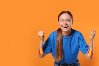 Image of Excited fan with colors matching Spain flag painted on her arms against orange background. Space for text
