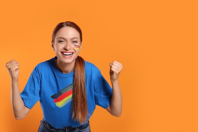 Image of Excited fan with flags of Germany on her cheek and t-shirt against orange background. Space for text
