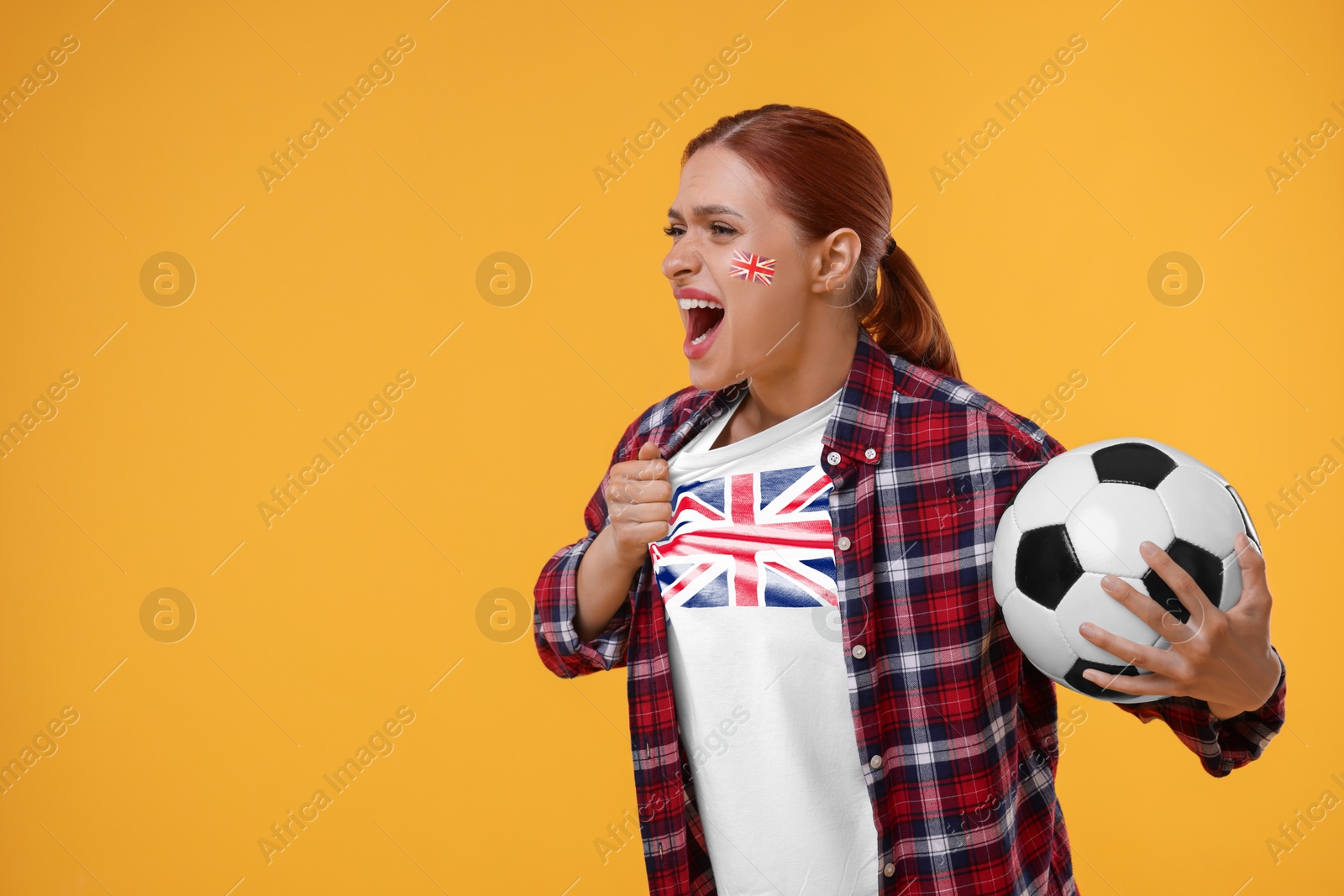 Excited fan with flags of United Kingdom on her cheek and t-shirt holding soccer ball against orange background. Space for text Image of Excited fan with flags of United Kingdom on her cheek and t-shirt holding soccer ball against orange background. Space for text