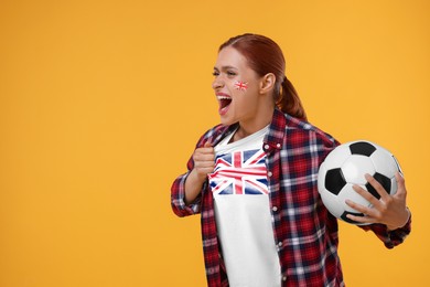 Image of Excited fan with flags of United Kingdom on her cheek and t-shirt holding soccer ball against orange background. Space for text