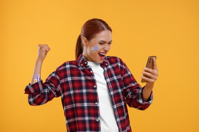 Image of Excited fan with flags of Uruguay painted on her body holding mobile phone against orange background. Celebrating victory