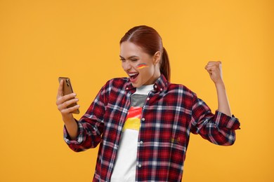 Image of Excited fan with flags of Germany on her cheek and t-shirt holding mobile phone against orange background. Celebrating victory