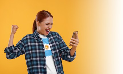 Image of Excited fan with flag of Argentina on her t-shirt holding mobile phone against orange background. Celebrating victory