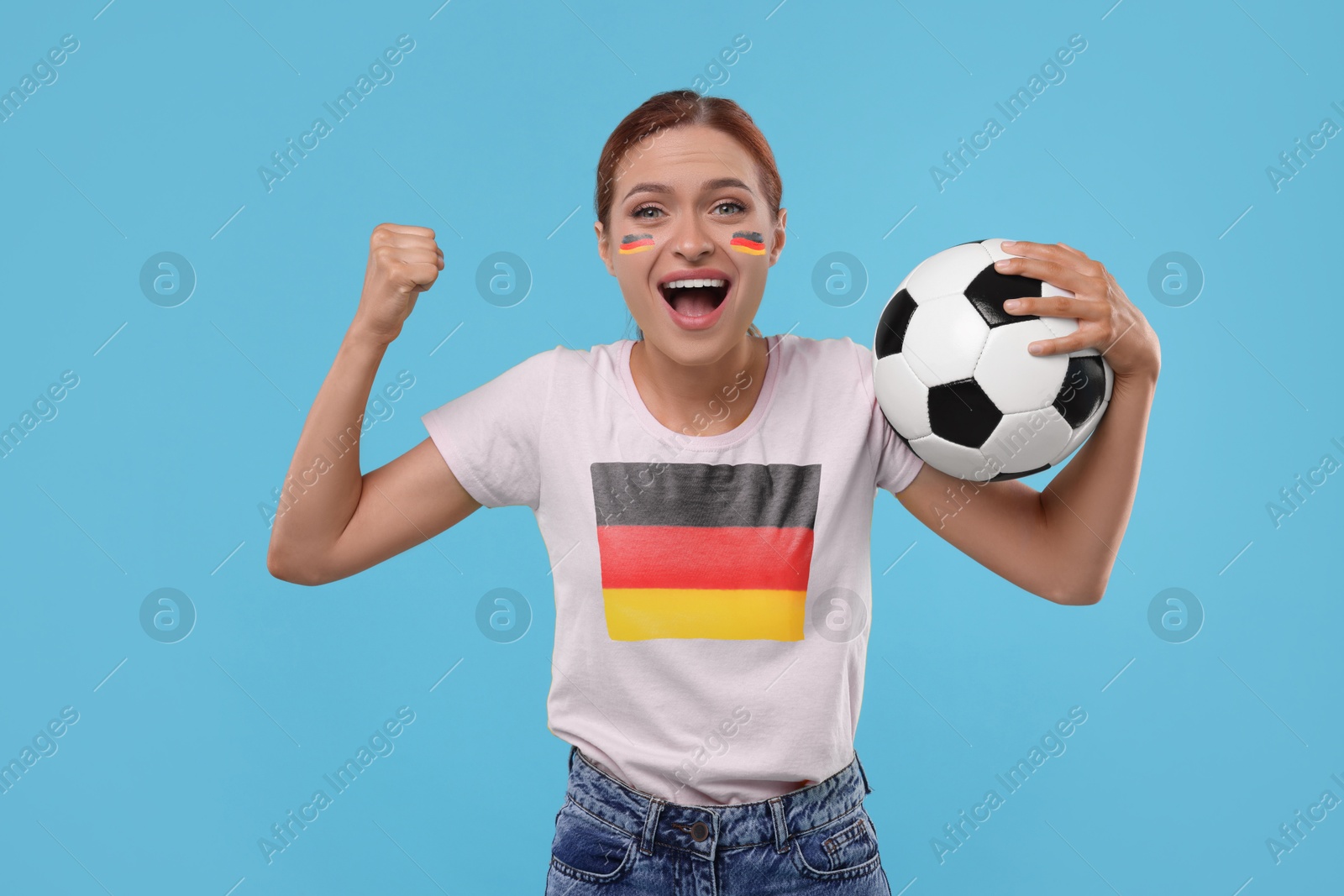 Excited fan with flags of Germany on her face and t-shirt holding soccer ball against light blue background Image of Excited fan with flags of Germany on her face and t-shirt holding soccer ball against light blue background