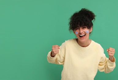 Image of Excited fan with flags of Uruguay painted on her face against green background. Space for text