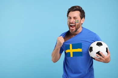 Image of Excited fan with flags of Sweden on his face and t-shirt holding soccer ball against light blue background. Space for text