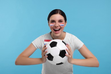 Image of Excited fan with flags of Netherlands on her face and t-shirt holding soccer ball against light blue background