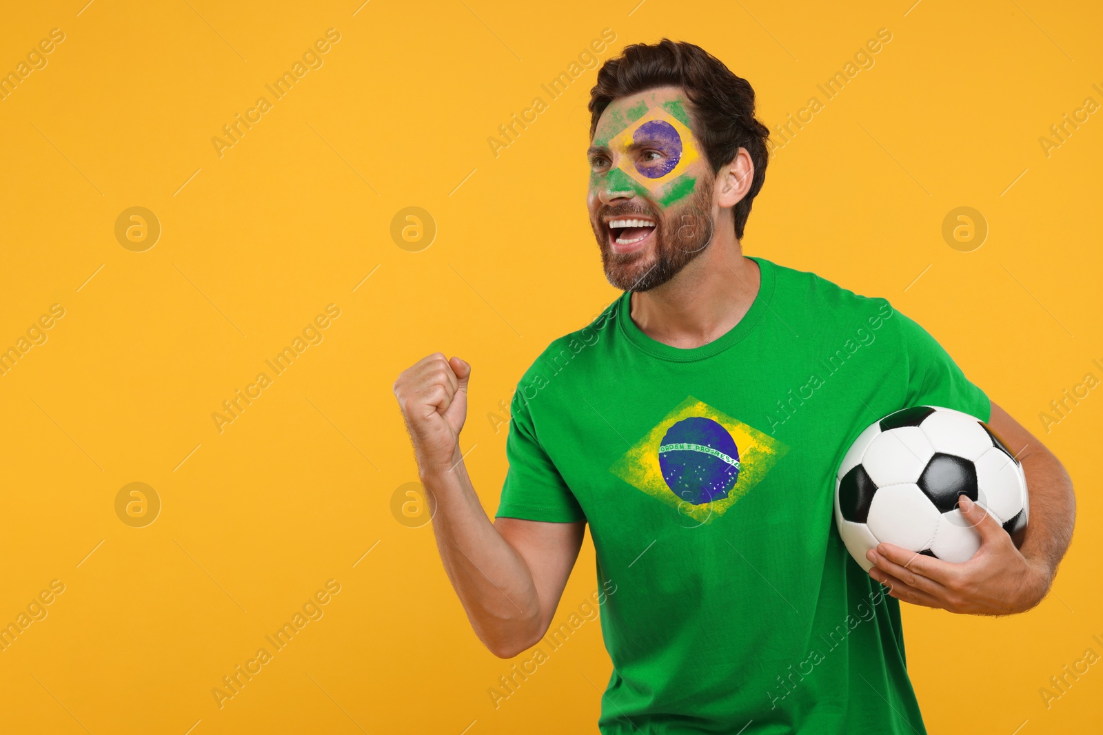 Excited fan with flags of Brazil on his face and t-shirt holding soccer ball against orange background. Space for text Image of Excited fan with flags of Brazil on his face and t-shirt holding soccer ball against orange background. Space for text