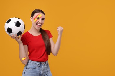 Image of Excited fan with colors matching Spain flag on her face and arm holding soccer ball against orange background. Space for text