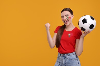 Image of Excited fan with flag of Portugal on her t-shirt holding soccer ball against orange background. Space for text