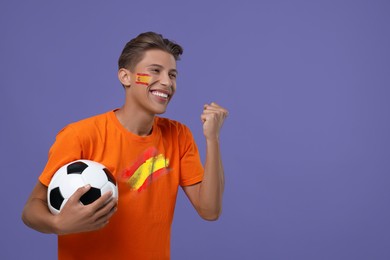 Image of Excited fan with flag of Spain on his face holding soccer ball against dark violet background. Space for text