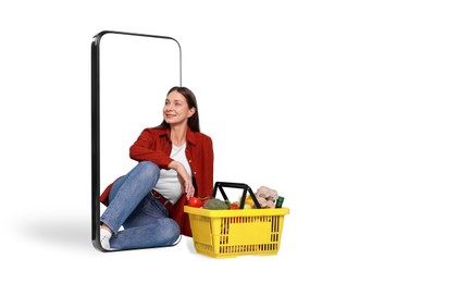 Image of Woman with shopping basket full of products sitting in mobile phone against white background. Online ordering