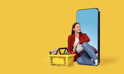 Image of Woman with shopping basket full of products sitting in mobile phone against orange background. Online ordering