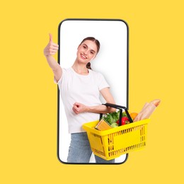 Image of Online ordering. Woman with shopping basket full of products showing thumbs-up out of mobile phone against orange background