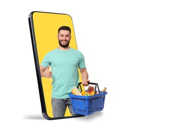 Image of Man with shopping basket full of products showing thumbs-up out of mobile phone against white background. Online ordering