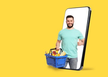 Image of Man with shopping basket full of products showing thumbs-up out of mobile phone against orange background. Online ordering