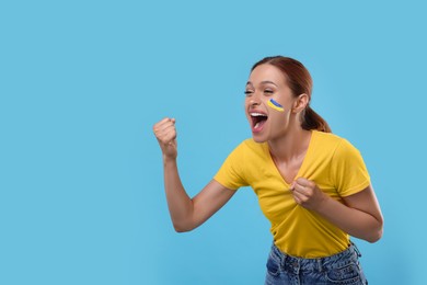 Image of Excited fan with flag of Ukraine painted on her cheek against light blue background. Space for text