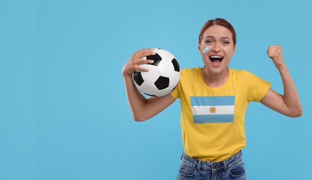 Image of Excited fan with flags of Argentina on her cheek and t-shirt holding soccer ball against light blue background. Banner design with space for text