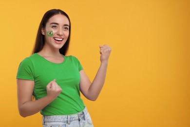Image of Excited fan with flag of Brazil painted on her cheek against orange background. Space for text