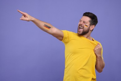 Image of Excited fan with flags of Germany painted on his cheek and arm against dark violet background. Space for text