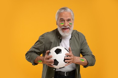 Image of Fan with flag of Brazil painted on his face holding soccer ball against orange background