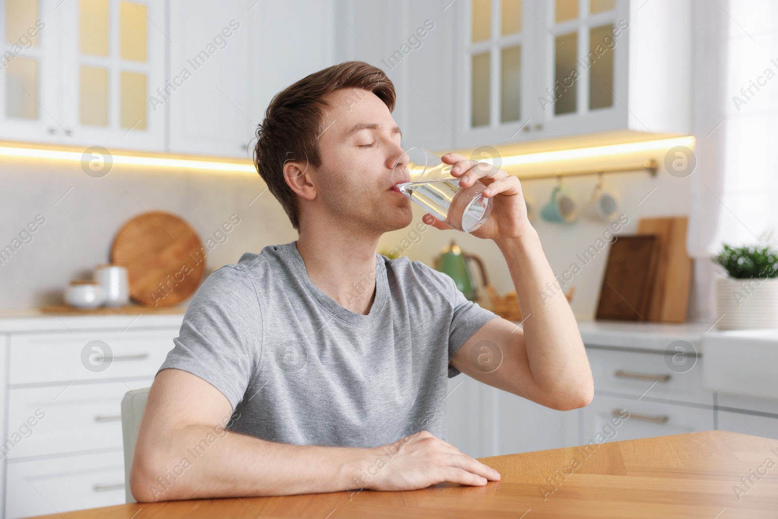 Photo of Man drinking water from glass at wooden table in kitchen
