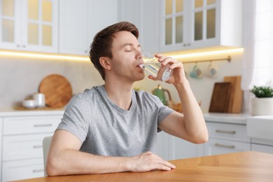 Photo of Man drinking water from glass at wooden table in kitchen