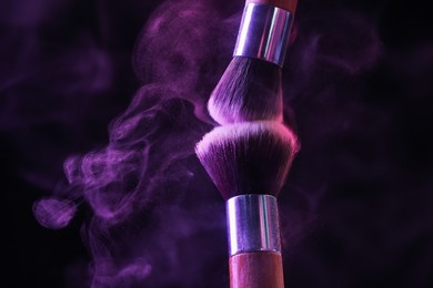 Photo of Makeup brushes with purple eye shadow in neon lights on black background, closeup