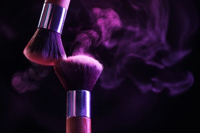 Photo of Makeup brushes with purple eye shadow in neon lights on black background, closeup. Space for text