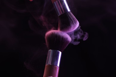 Photo of Makeup brushes with purple eye shadow in neon lights on black background, closeup
