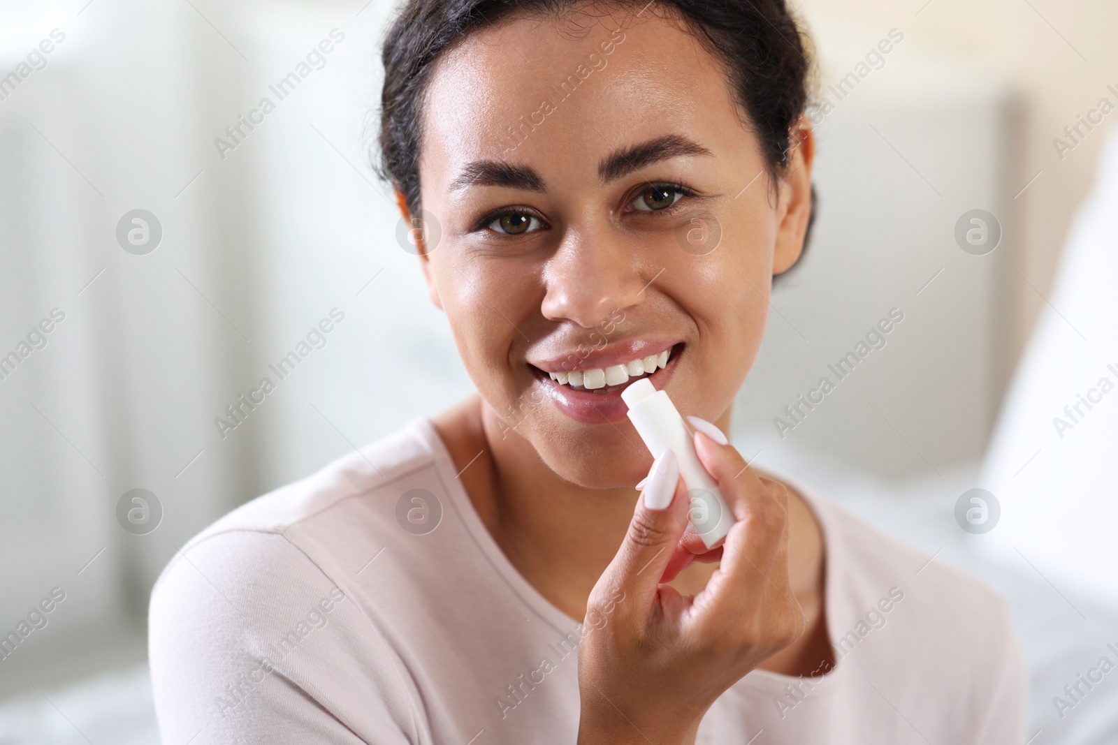 Photo of Smiling woman applying lip balm onto her lips on blurred background, closeup