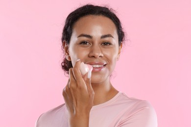 Photo of Smiling woman applying lip balm onto her lips on pink background