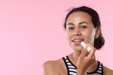 Photo of Smiling woman applying lip balm onto her lips on pink background. Space for text