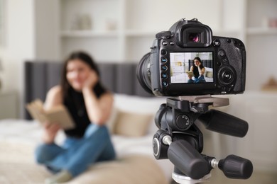 Photo of Woman reading book on bed in studio, focus on video camera recording her