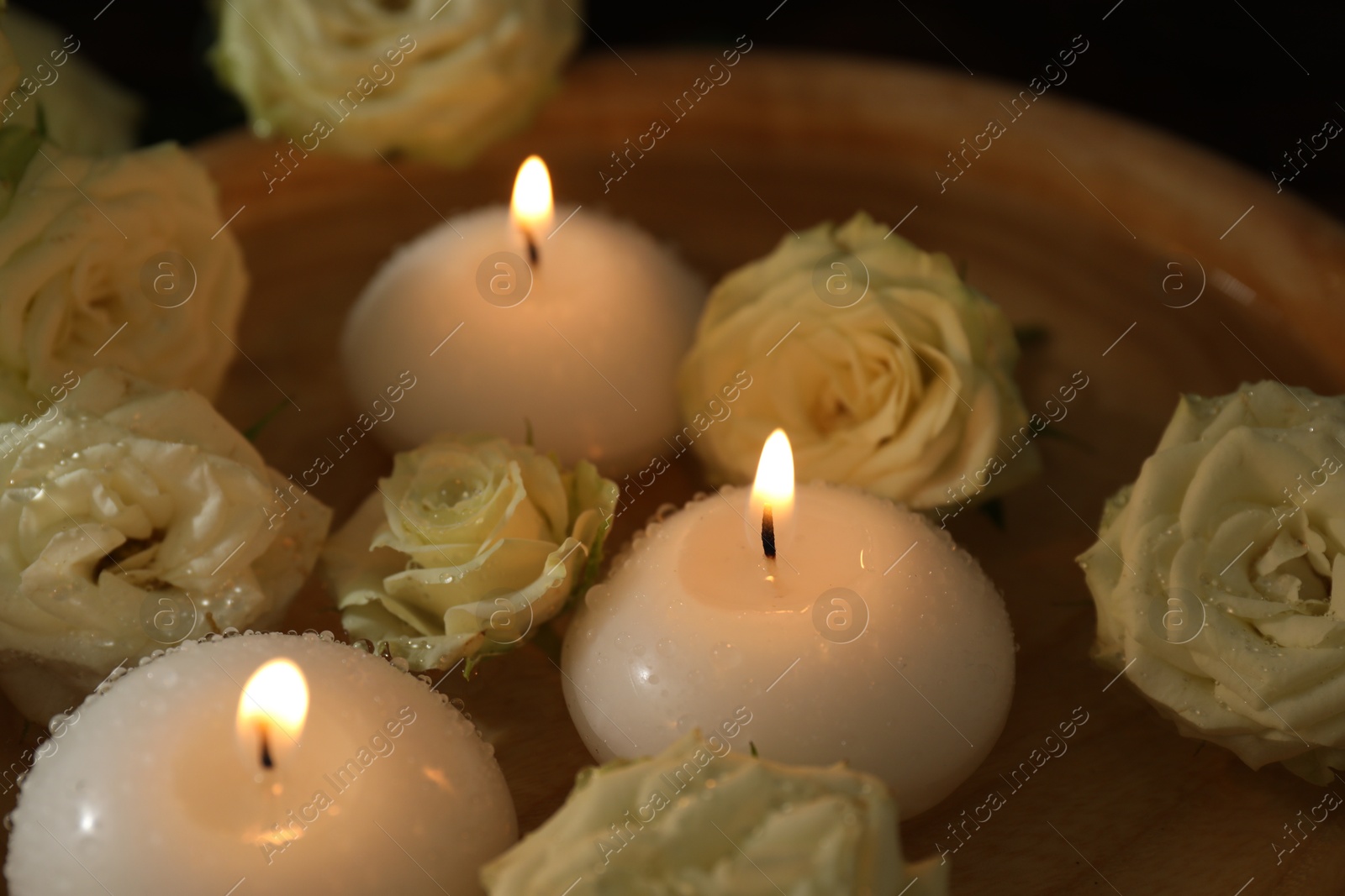 Photo of Burning candles, water and rose flowers in wooden bowl on table, closeup