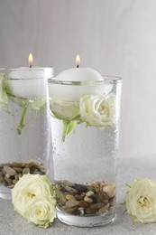 Photo of Burning candles in glasses with water, stones and rose flowers on light grey table, closeup