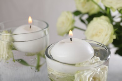 Photo of Burning candles in glasses with water and rose flowers on table, closeup