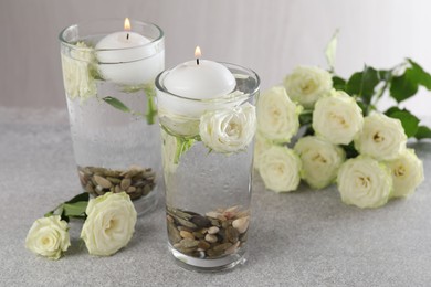 Photo of Burning candles in glasses with water, stones and rose flowers on light grey table, closeup