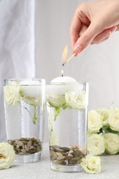 Photo of Woman lighting candles in glasses of water at light grey table, closeup