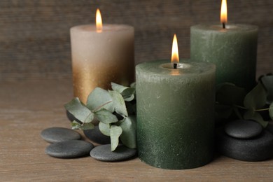 Photo of Burning candles, stones and eucalyptus branches on wooden table, closeup